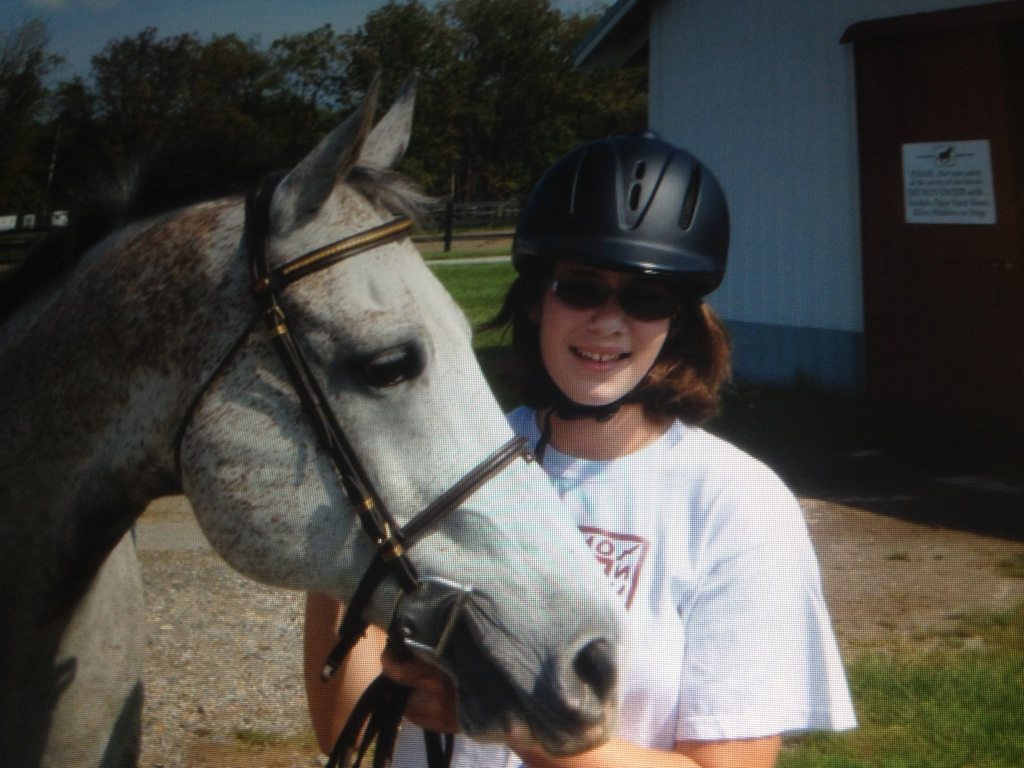 A close-up view shows a young rider standing holding a horse tacked up in English gear, the head and neck of the horse visible. The horse is primarily a light grey color, but his coat displays intricate variations in shading and patterns. Across the face and neck, there are dapples and speckles in darker grey, giving it a textured, slightly mottled appearance. His muzzle and around his eyes are a slightly deeper, charcoal-toned grey compared to the rest of his face. His mane has a mix of both black and gray strands, with darker hair along the top. Around the lower jaw and under the chin, the grey becomes subtly lighter, almost merging into white in some places. Some of the flex of color and dapples have a more reddish brown hue than the rest of the coat. 
